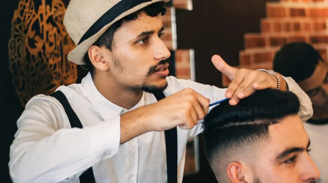 A barber in a straw trilby combing a client's hair against a brick wall